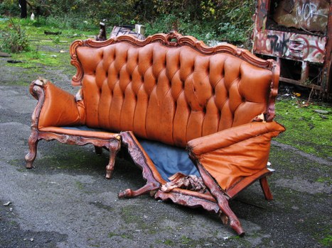 A distressed antique sofa with a wooden carved frame and tufted orange upholstery, showing significant damage with the cushions and fabric torn and hanging loose, placed outdoors on a rough asphalt surface surrounded by green grass and trees in the background. The environment appears to be a waste disposal or rubbish collection area, with parts of the sofa indicating it is no longer functional. This scene is typical of furniture waste collected by a rubbish removal service, such as Commercial Waste Chesham, in the Chesham area of Buckinghamshire. The overall setting suggests the sofa has been discarded and is awaiting removal, fitting within the context of waste disposal or clearance services specializing in furniture collection in the local vicinity.