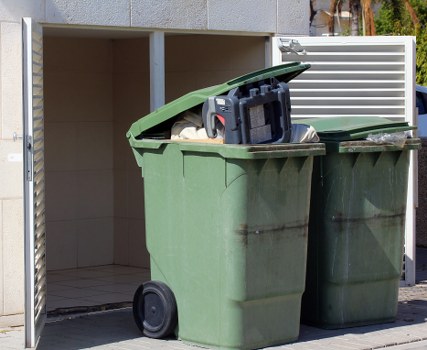 The image shows two large green wheeled rubbish bins placed outside a building, positioned on a paved surface. The first bin's lid is open, revealing mixed waste, including plastic bags and cardboard. The second bin, with its lid closed, is situated beside the open one. Behind the bins, part of a white slatted fence is visible, and an area of beige tiles or wall can be seen inside an open space or storage shed. The environment appears to be an outdoor area, possibly a driveway or back yard, with natural daylight illuminating the scene. The presence of commercial waste bins aligns with rubbish removal services, such as those offered by Commercial Waste Chesham, operating within the Chesham postcode area.