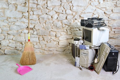 In a room with a textured stone wall background, a wooden broom with a yellow handle leans upright against the wall on the left side of the image. On the floor in front of the broom, there is a small pink plastic dustpan with a short handle. To the right of the scene, a collection of electronic waste and miscellaneous items are stacked and scattered, including an old CRT monitor, a desktop computer tower, a keyboard, a mouse, and various cables and power supplies. Additionally, there's a vintage-style radio and a black speaker positioned at the bottom right corner. The area appears to be a designated space for collecting or storing waste items, with a concrete or cement floor, indicative of an indoor environment or a garage setting, aligned with the rubbish removal services offered by Commercial Waste Chesham in the Chesham area.