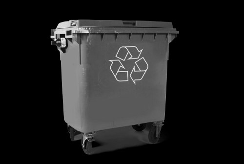 A standard black plastic wheeled rubbish bin with a hinged lid, marked with a white recycling symbol on the front, is positioned on a concrete driveway. The bin's surface has a matte finish and appears clean without visible dirt or damage. The background is plain and dark, emphasizing the bin’s shape and features. The bin is situated in an outdoor setting typical for waste collection, possibly near a property or commercial premises in Chesham. The scene captures the bin ready for waste disposal or collection, aligning with services provided by Commercial Waste Chesham for efficient rubbish removal in the local area.