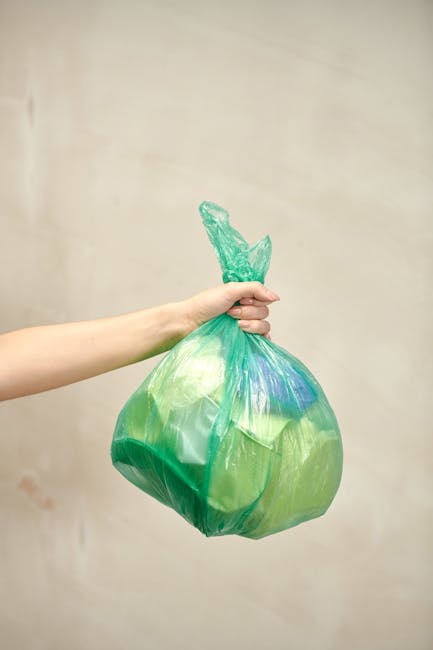 A large pile of clear plastic rubbish bags filled with assorted waste, including clothing, paper, and other household items, is situated on a paved sidewalk outside a commercial store. Several black a